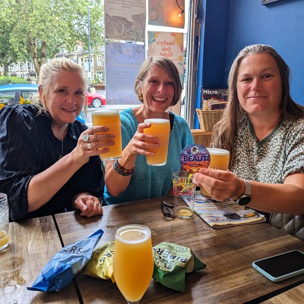 Three friends raising pints of hazy pale at a window table, one holding up the Beaut pump clip